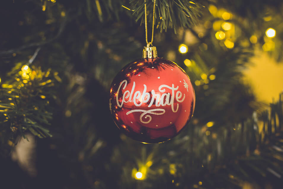 Esfera roja de Navidad, con la palabra "Celebrate", colgada en árbol de Navidad con luces