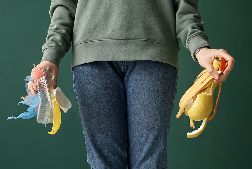 Persona con suéter verde y jeans azules, sosteniendo con su mano derecha residuos inorgánicos, y con su mano izquierda residuos orgánicos