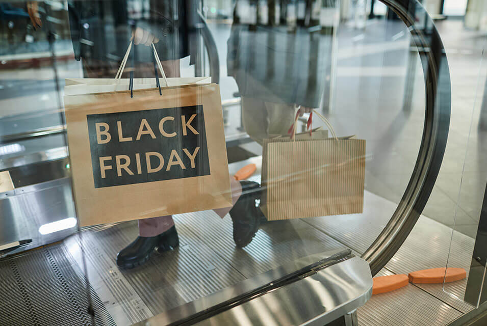 Black Friday escrito en bolsa de compras, sostenida por persona mientras desciende por escaleras eléctricas dentro de centro comercial