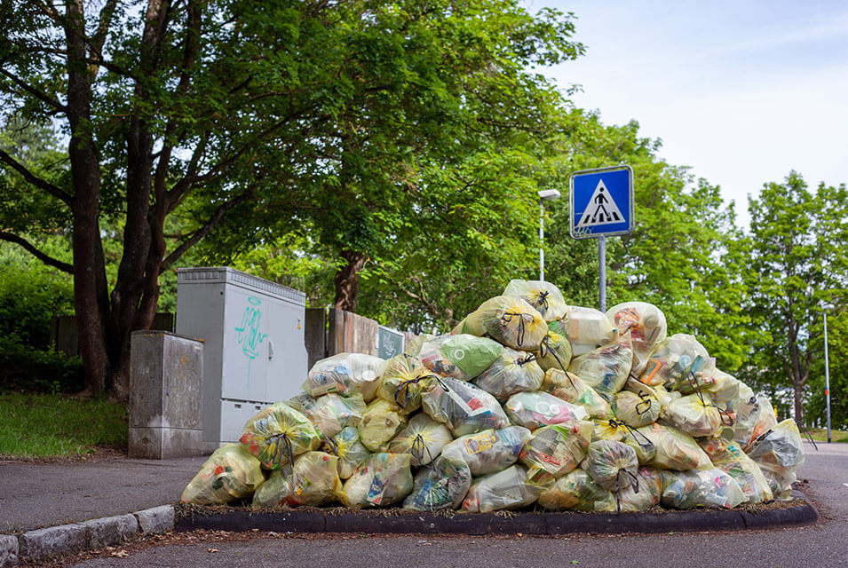 Bolsas de basura apiladas sobre pavimento de la calle, con árboles, señal de tránsito y paredes grises, al fondo, haciendo referencia a la diferencia entre residuo y basura